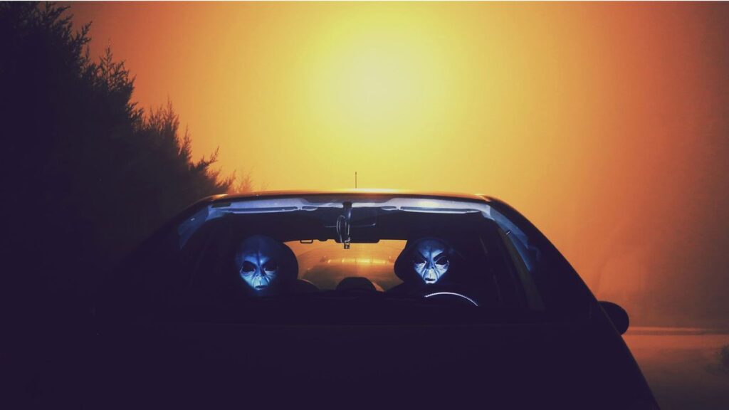 Eerie alien figures with masks seen driving a car during a colorful sunset.