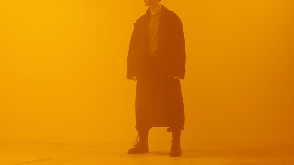 Man in black coat standing against an orange background, embodying a cyberpunk retro-futuristic style.