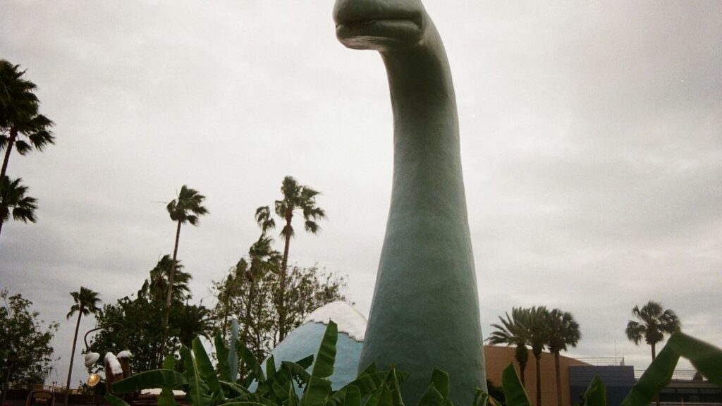 A large dinosaur statue surrounded by palm trees under a cloudy sky.