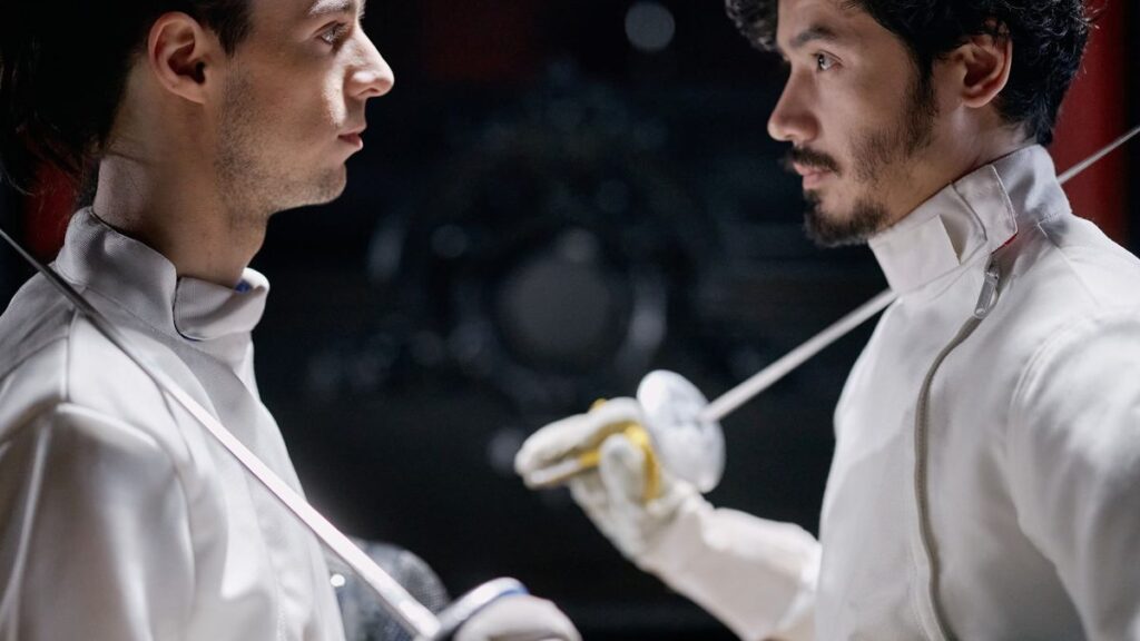 Close-up of two male fencers in action, facing each other with foils ready in a dimly lit salle.